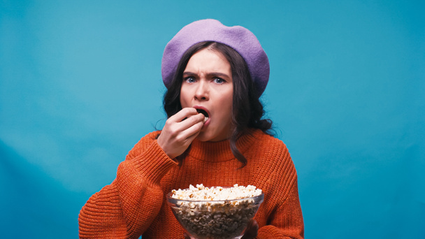 worried woman in lilac beret eating popcorn while watching movie isolated on blue - Photo, Image