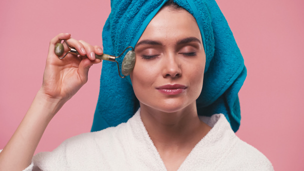 young woman with towel on head massaging face with jade roller isolated on pink - Photo, Image