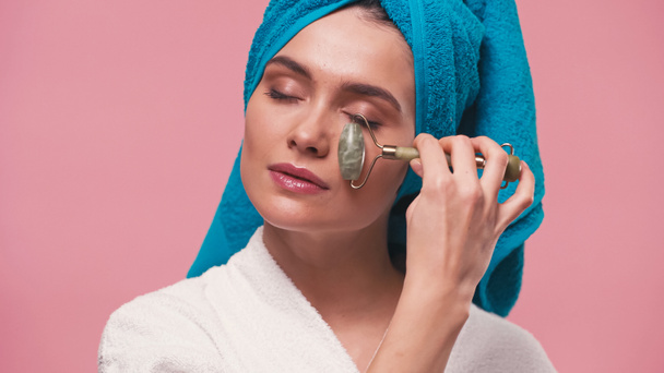 young woman with towel on head and closed eyes massaging face with jade roller isolated on pink - Φωτογραφία, εικόνα