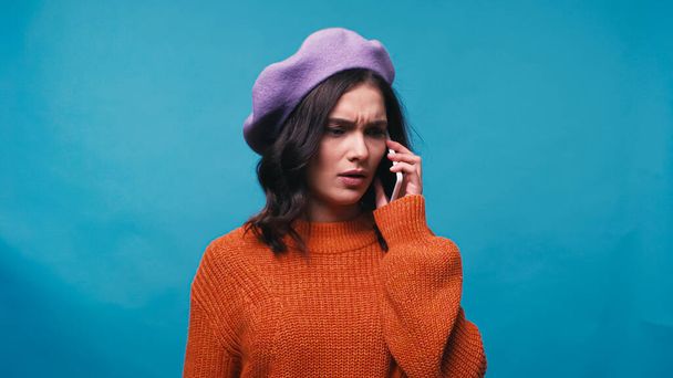 thoughtful woman in beret talking on smartphone isolated on blue - Fotoğraf, Görsel