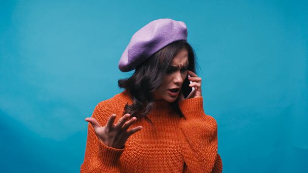 worried woman gesturing while talking on mobile phone isolated on blue - Foto, Imagen