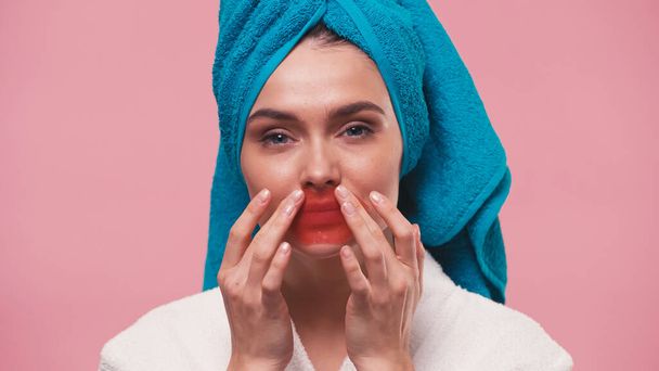 young woman with towel on head looking at camera and applying lip mask isolated on pink - Photo, Image