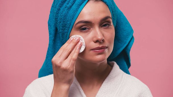 woman with terry towel on head wiping face with cotton pad isolated on pink - Foto, afbeelding