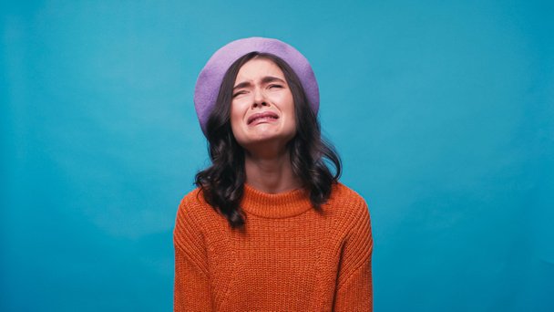sad woman in warm jumper and beret crying isolated on blue - Foto, imagen