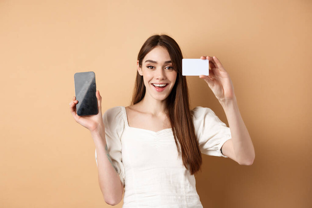 Joyeux jeune femme montrant carte de crédit en plastique et écran de téléphone portable vide, souriant heureux à la caméra, debout sur fond beige - Photo, image