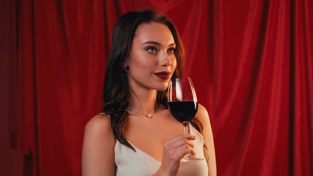 young woman holding glass of wine and smiling on red - Фото, изображение