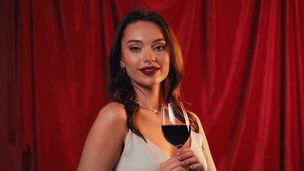 positive young woman holding glass of wine on red - Foto, imagen