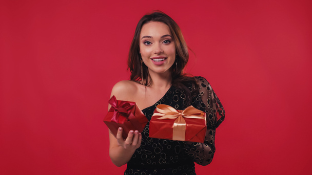 cheerful young woman smiling and holding gift boxes isolated on red - Фото, зображення