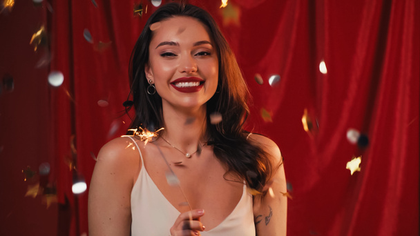 cheerful woman with tattoo holding bright sparkler near confetti on red - Photo, Image