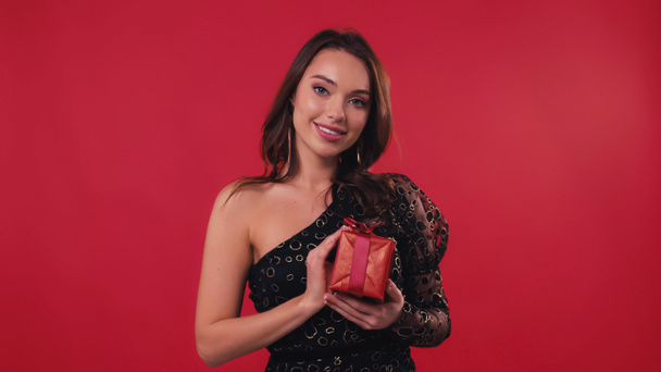 joyful young woman smiling while holding gift box isolated on red - Fotoğraf, Görsel