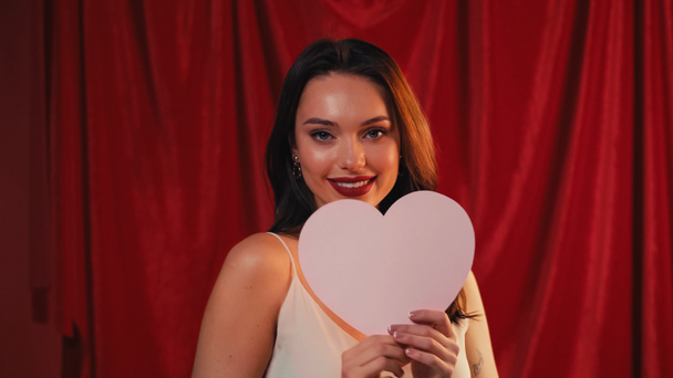 positive woman holding paper heart and smiling on red - 写真・画像
