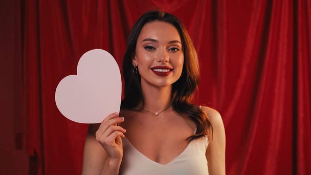 joyful woman holding paper heart and smiling on red - 写真・画像