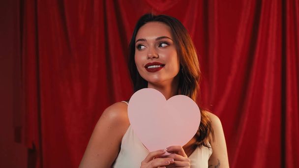 happy woman holding paper heart and looking away on red - Foto, imagen