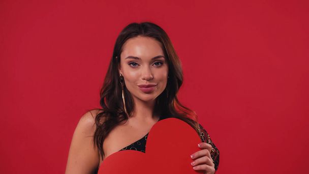 young and pleased woman holding paper heart isolated on red - Photo, Image