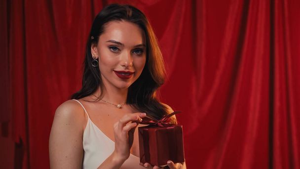 elegant woman smiling and holding gift box on red - Fotoğraf, Görsel
