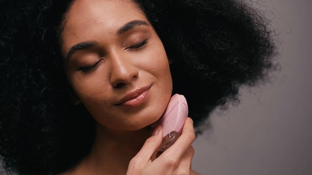 piacere donna afroamericana utilizzando detergente siliconico sul viso isolato su grigio - Foto, immagini