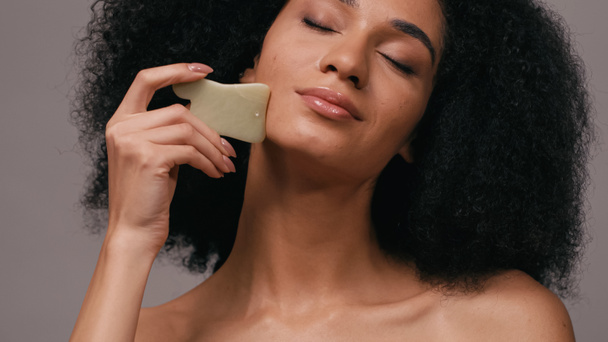 african american woman with closed eyes using gua sha stone isolated on grey - Foto, Imagem