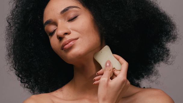brunette african american woman with closed eyes using gua sha stone isolated on grey - Foto, Bild