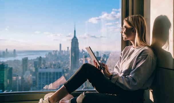 Side view of happy millennial digital nomad with modern touch pad looking in lookout window and dreaming about New York sightseeing, carefree hipster girl enjoying time in high rise building - Фото, изображение