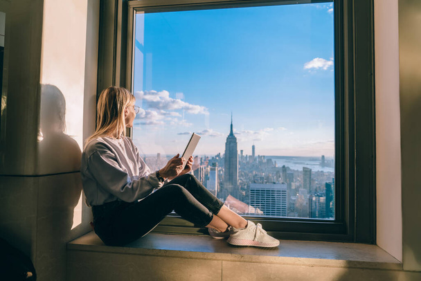Thoughtful female freelancer with modern touch pad in hands looking in panoramic window with beautiful scenery of New York megalopolis, millennial woman with digital tablet dreaming about travel - Фото, зображення