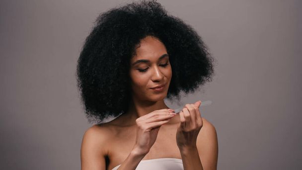 кудрявая молодая африканская американка, использующая пилку для ногтей, изолированную на серой - Фото, изображение