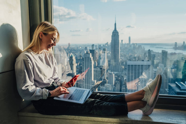 Happy millennial woman browsing influence website during travel in New York working with media files in high rise with panoramic views on Manhattan landscape, cheerful blogger watching video - Foto, Imagem