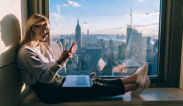 Millennial distance worker sharing multimedia files via cellphone and laptop technology connecting to bluetooth, skilled freelancer chatting on smartphone sitting at panoramic window in New York - Valokuva, kuva