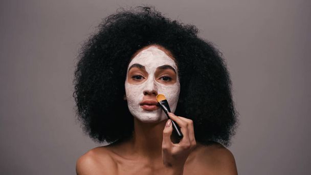 кудрявая африканская американка с голыми плечами надевает глиняную маску, изолированную на сером - Фото, изображение
