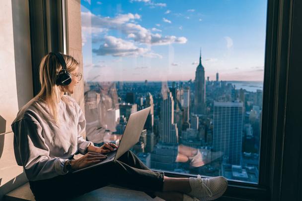 Millennial female freelancer in electronic headphones for noise cancellation listening positive music podcast and looking in panoramic window with breathtaking Manhattan view from high rise - Foto, imagen