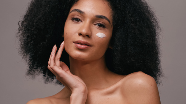 curly african american woman with cosmetic cream on face isolated on grey - Фото, зображення