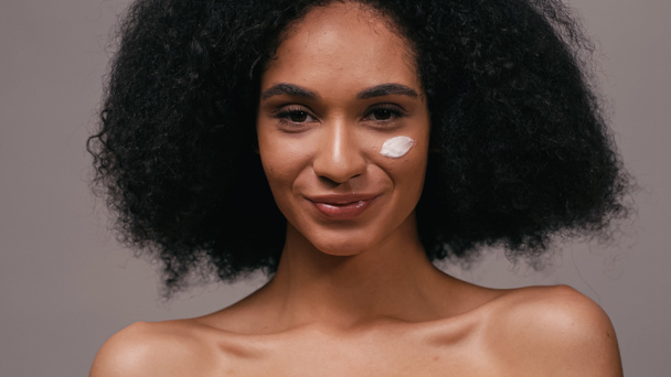 donna afroamericana sorridente con crema cosmetica sul viso isolata sul grigio - Foto, immagini