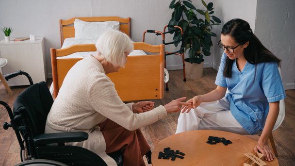 senior femme en fauteuil roulant jouer dominos avec brunette infirmière - Photo, image