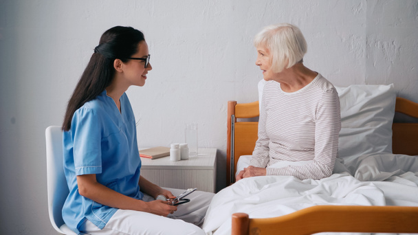 brunette verpleegster praten met gelukkige oude patiënt en het houden van stethoscoop  - Foto, afbeelding