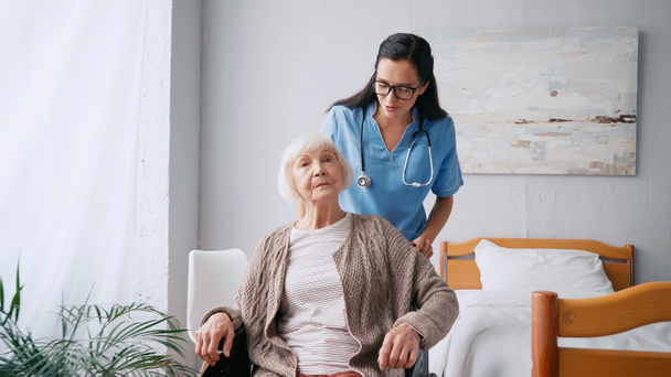 brunette nurse in eyeglasses moving aged woman in wheelchair  - Photo, Image