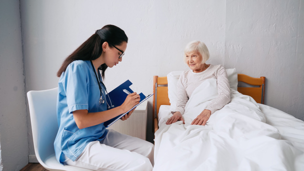brunette verpleegster in bril schrijven recept in klembord in de buurt van oude vrouw in bed - Foto, afbeelding