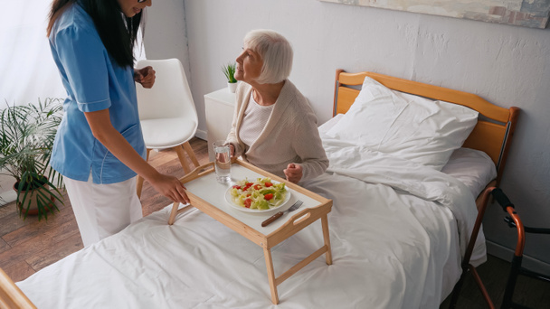 brunette nurse talking with aged patient near breakfast tray on bed  - Foto, afbeelding