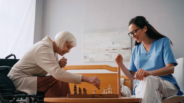 ευτυχισμένη ηλικιωμένη γυναίκα και χαμογελαστή νοσοκόμα που παίζει σκάκι στο γηροκομείο - Φωτογραφία, εικόνα