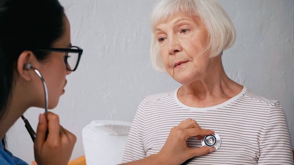 niewyraźna pielęgniarka w okularach badająca starszą kobietę stetoskopem  - Zdjęcie, obraz