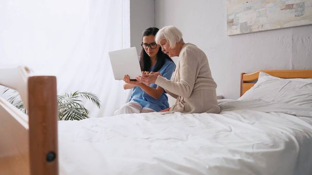 брюнетка пожилая медсестра показывает пожилой женщине, как использовать ноутбук в доме престарелых  - Фото, изображение
