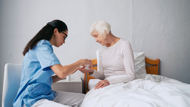 brunette verpleegster in bril het geven van pillen aan oudere vrouw in kliniek - Foto, afbeelding