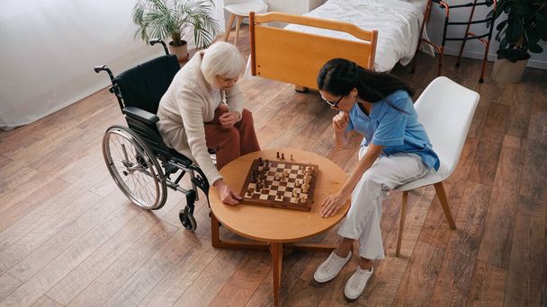 Huzurevinde satranç oynayan yaşlı kadın ve hemşirelere yüksek açılı bakış açısı - Fotoğraf, Görsel