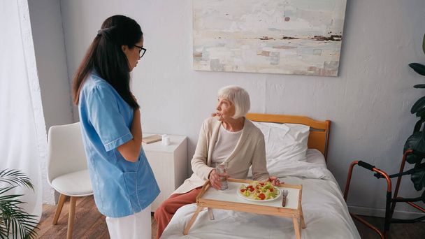 enfermera morena hablando con paciente anciano cerca de bandeja con desayuno en la cama  - Foto, imagen