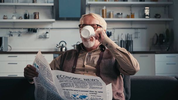 Senior man in eyeglasses holding newspaper while drinking tea at home - Photo, image