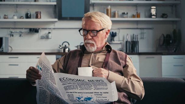 senior man in eyeglasses reading newspaper while holding cup of tea - Fotó, kép