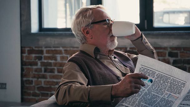szakállas idősebb férfi szemüvegben, aki újságot tart teázás közben.  - Fotó, kép