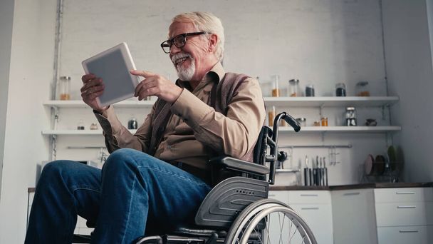 Glimlachende gehandicapte gepensioneerde in rolstoel met digitale tablet thuis - Foto, afbeelding