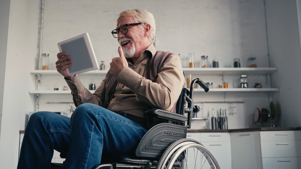 Pensionista discapacitado sonriente en silla de ruedas mostrando el pulgar hacia arriba durante la videollamada en casa - Foto, imagen