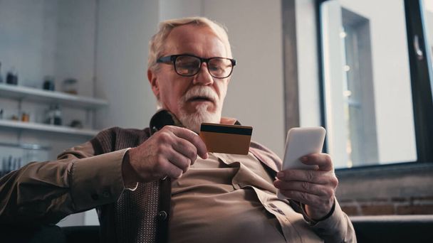 自宅でオンラインショッピングしながらクレジットカードを携帯電話を保持しているシニア男 - 写真・画像