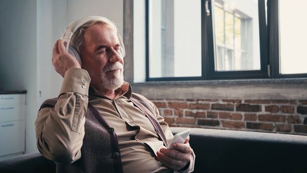 zadowolony i starszy mężczyzna w słuchawkach bezprzewodowych słuchanie muzyki i trzymanie smartfona w domu  - Zdjęcie, obraz