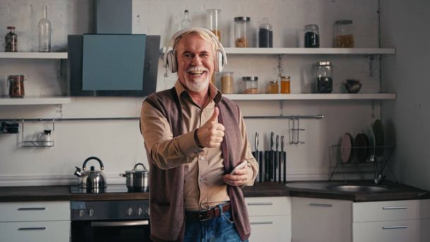 šťastný a starší muž v bezdrátových sluchátkách poslech hudby, držení smartphone a ukazující palec nahoru v kuchyni - Fotografie, Obrázek